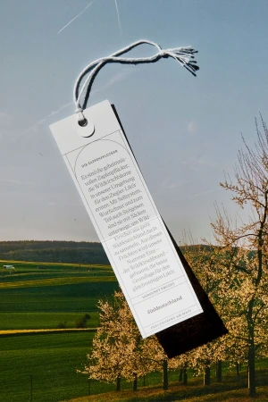 A vertical bookmark with text hangs in front of an outdoor scene of green fields and flowering trees under a bright blue sky.