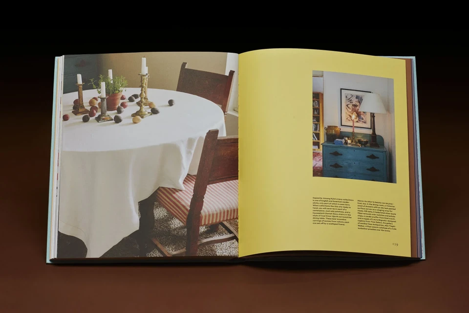 Interior design book features a round table with a white tablecloth, antique brass candlesticks, fruit, and a rustic wooden chair.