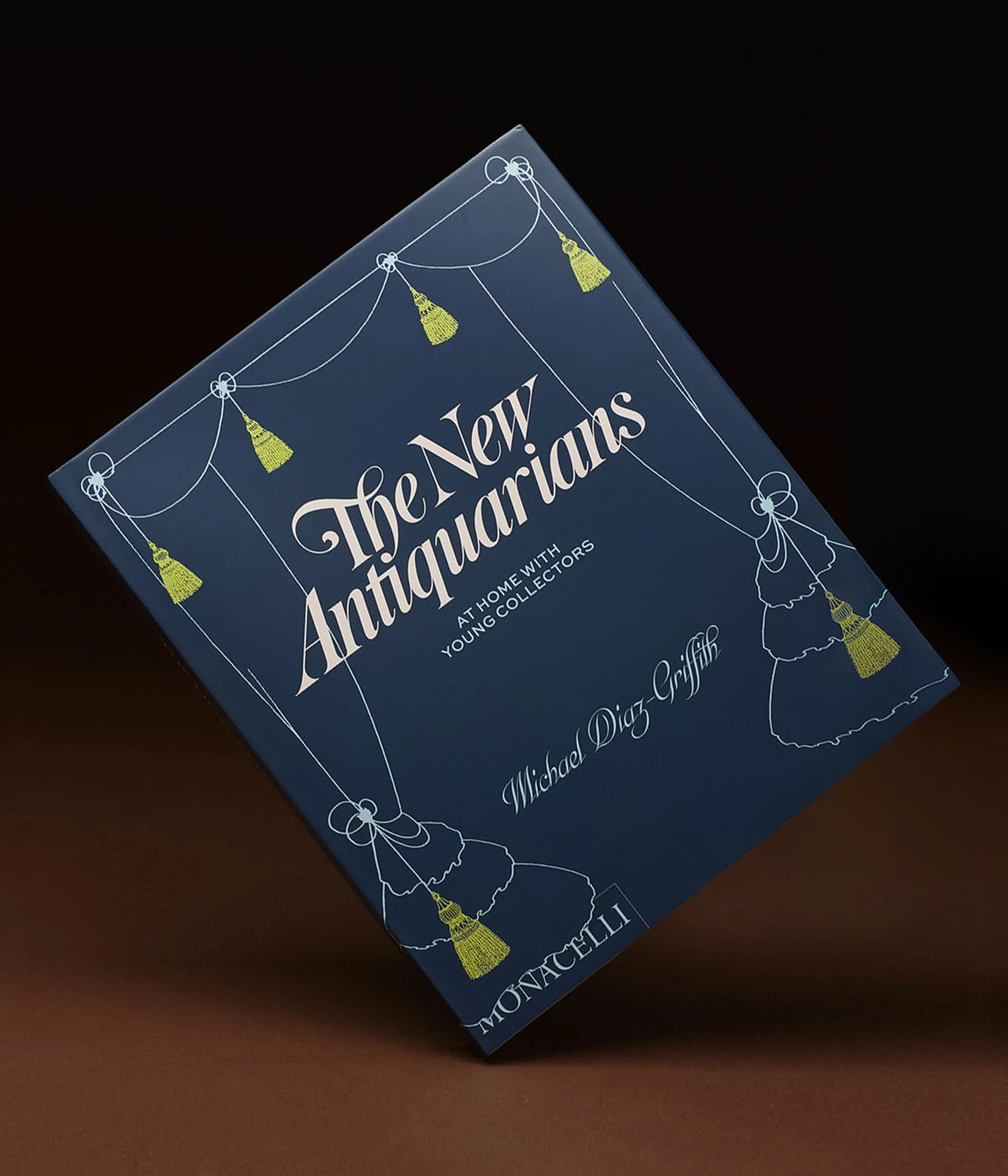 Blue hardcover book tilted on a brown surface features the title 'The New Antiquarians' with tassels and author information on the cover.