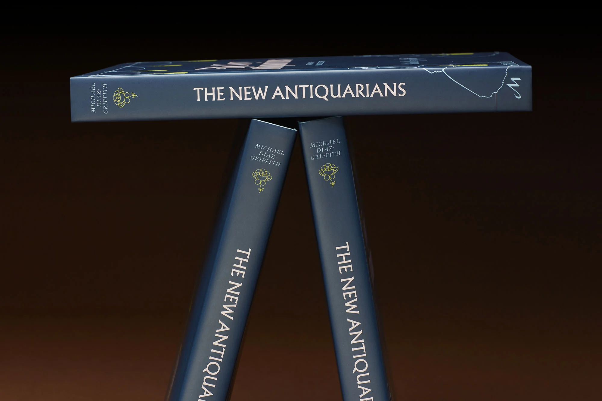 Three copies of the book 'The New Antiquarians' are stacked in a T-shape formation, against a dark brown background, showcasing book design.
