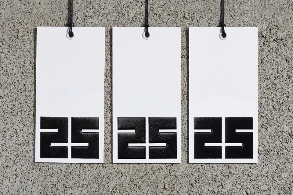 Three white tags hang against a textured gray wall, each featuring a black graphic design at the bottom, minimalist studio shot.