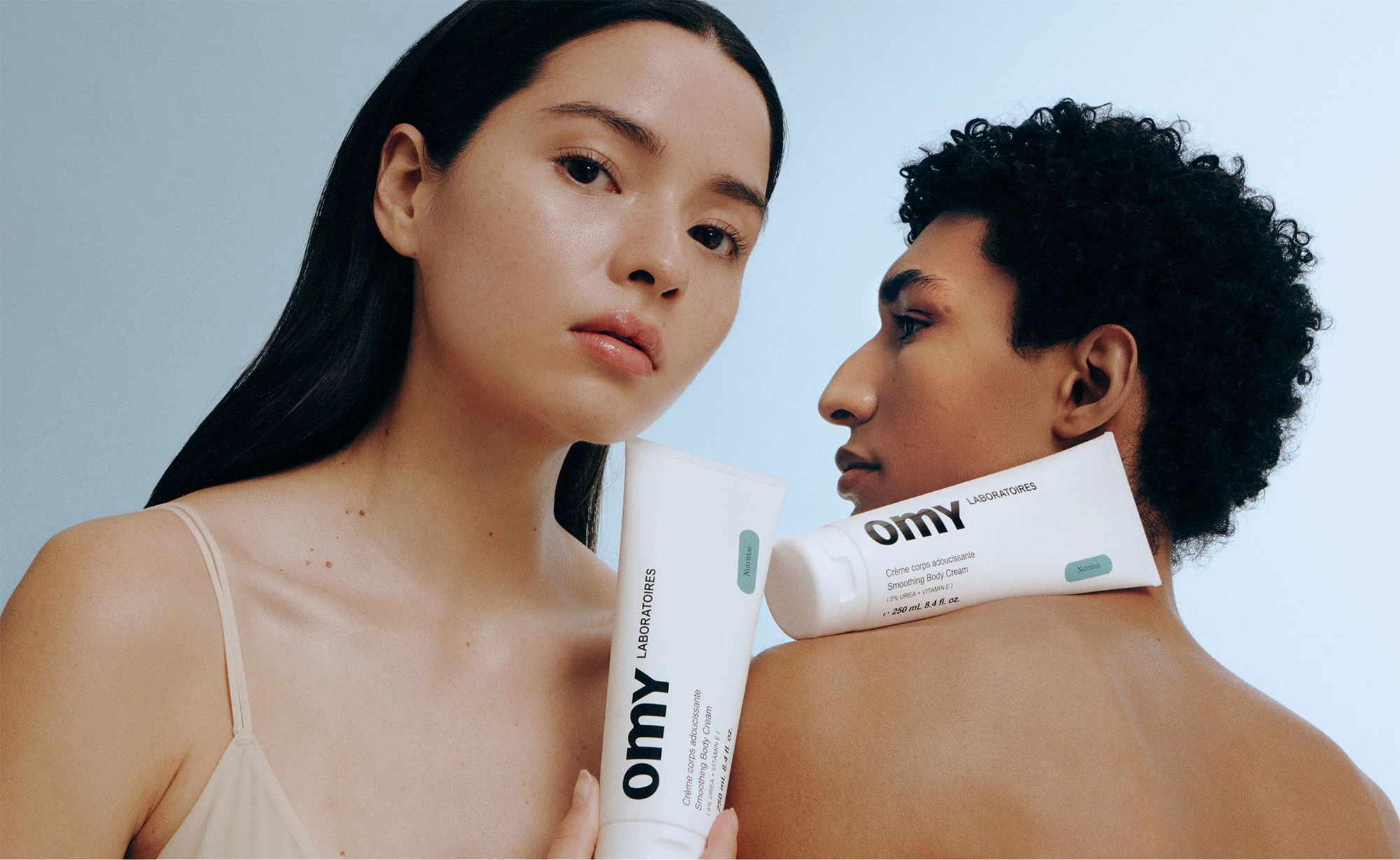 Two models, one male and one female, with Omy Laboratoires skincare products against a light blue background in a beauty shot.