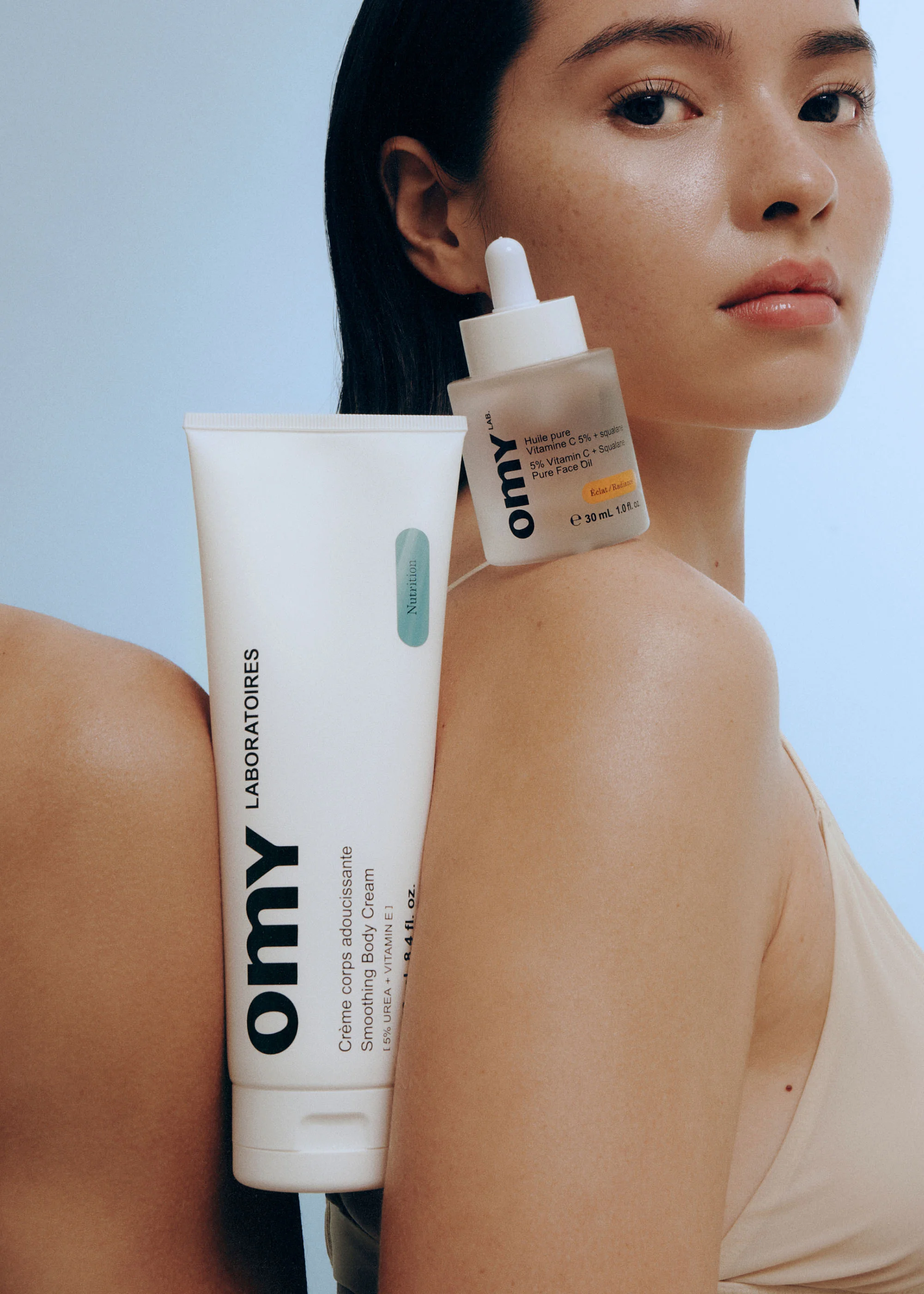 Asian woman poses with OMY skincare products: body cream tube and face serum, with a light blue background, promoting beauty and self-care.