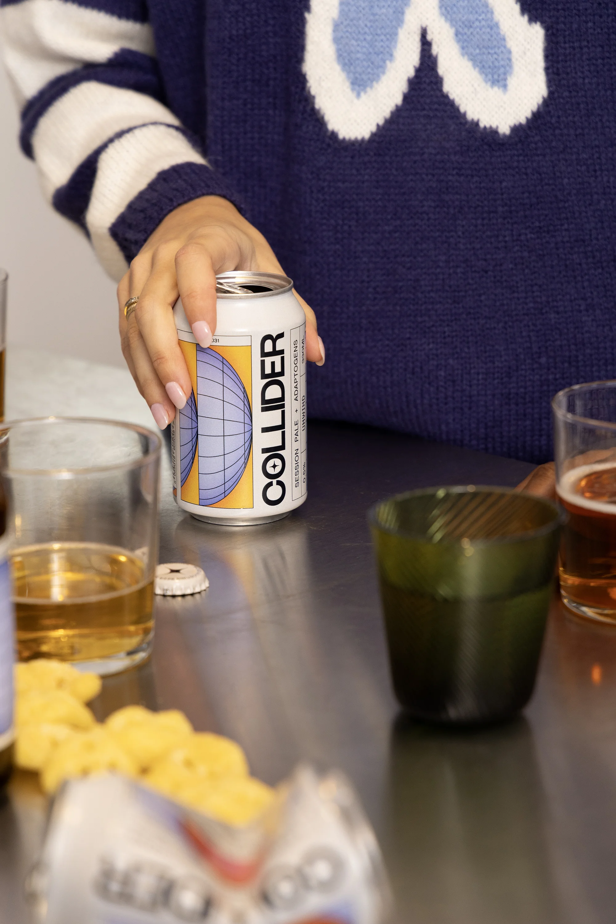 Close-up on hand holding a can of Collider beer on a metal table with drinks, glasses, and snack food in a casual indoor setting.
