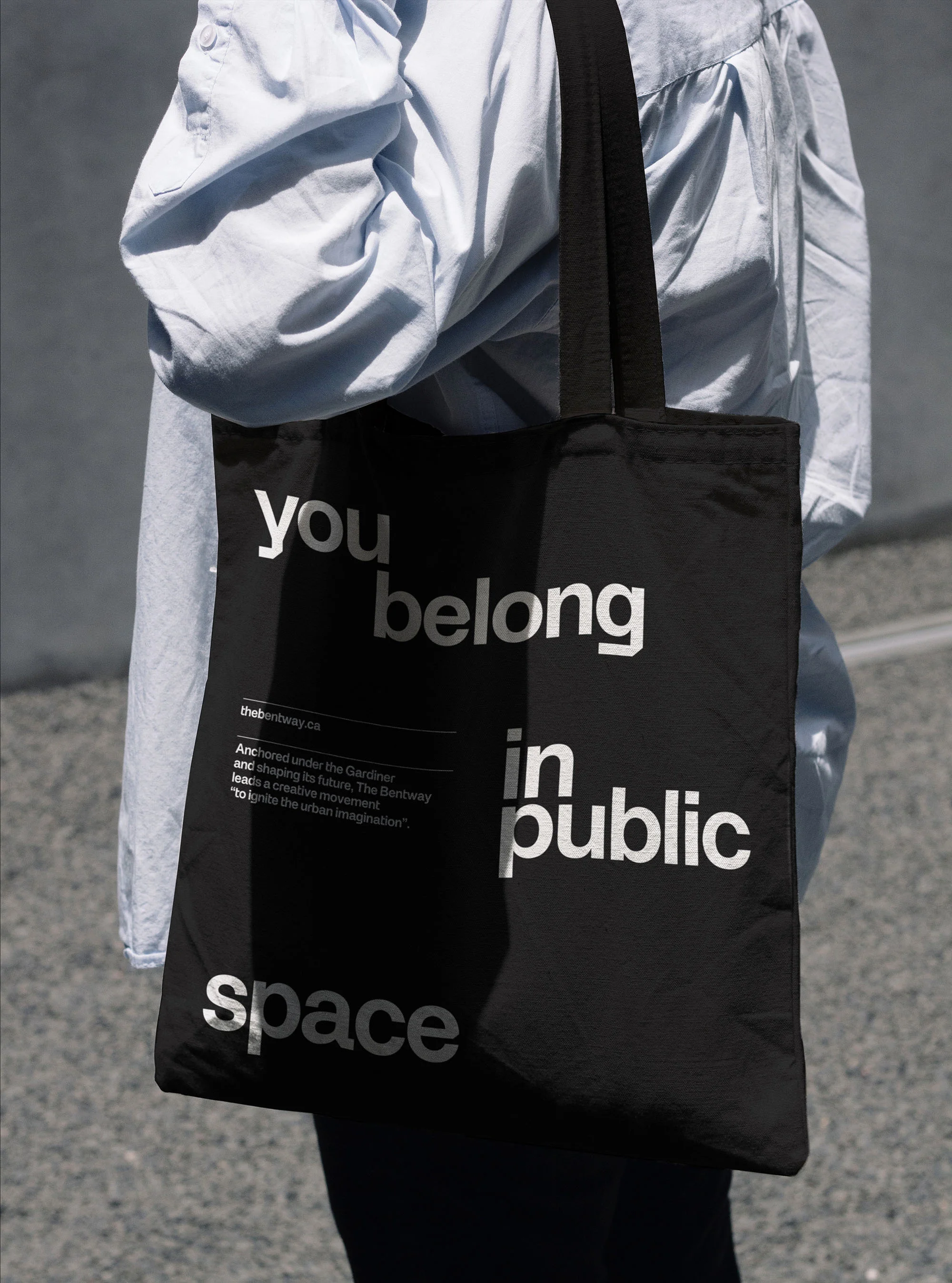 Black tote bag with white text 'You belong in public space' held by a person in a light blue shirt against a grey concrete background.