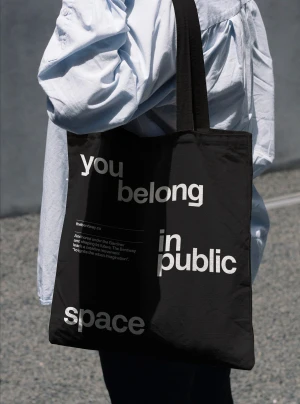 Person wearing a light blue button-down shirt carrying a black tote bag with white typographic text reading you belong in public space.