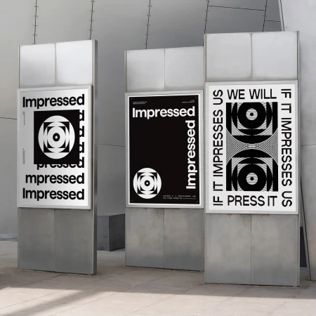Three posters on metal stands display the word "Impressed" in black and white designs with geometric patterns, set indoors on a gray floor.