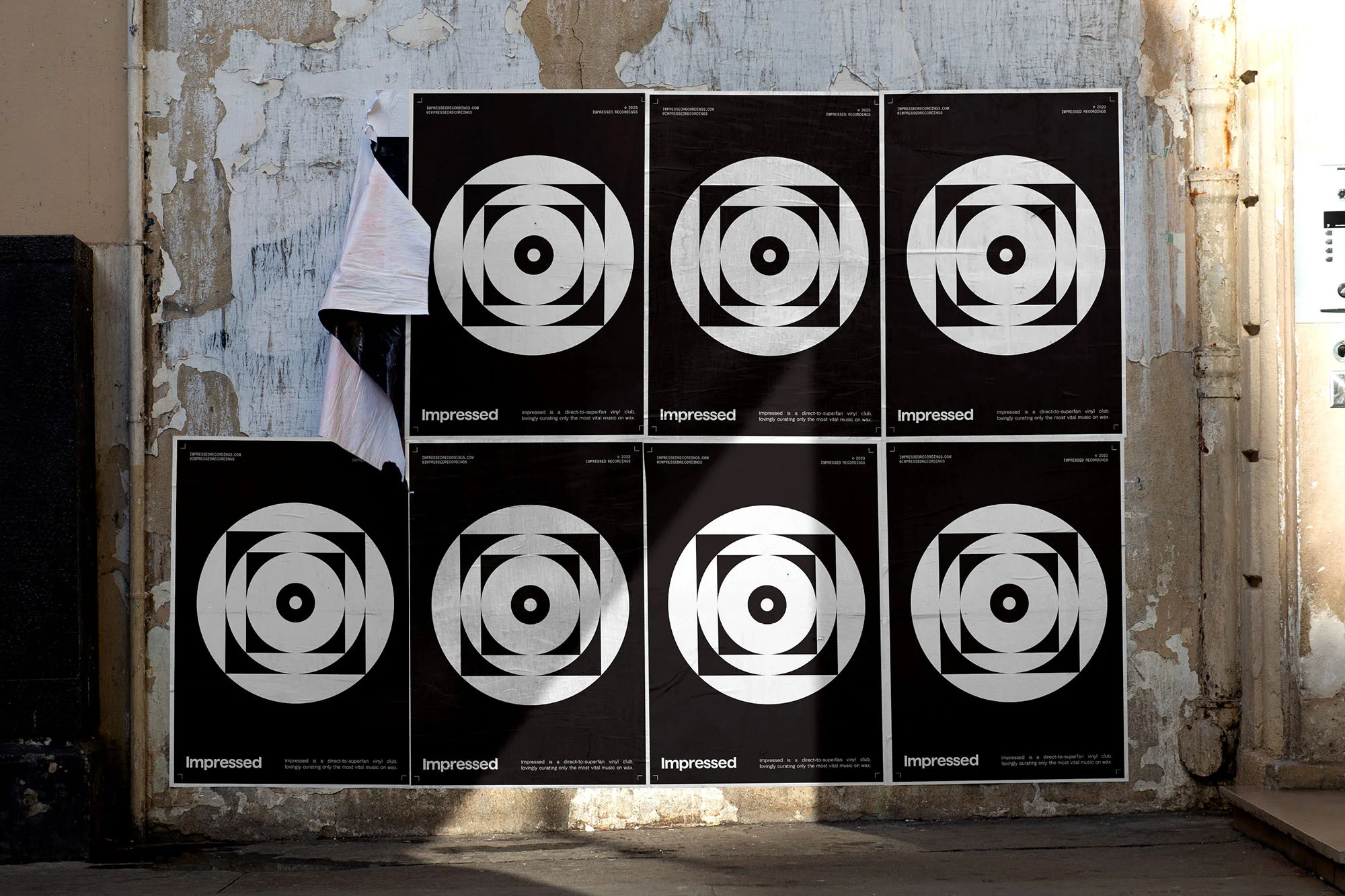 Multiple black posters with a concentric geometric circle and square design are adhered to an aged, textured wall indoors.