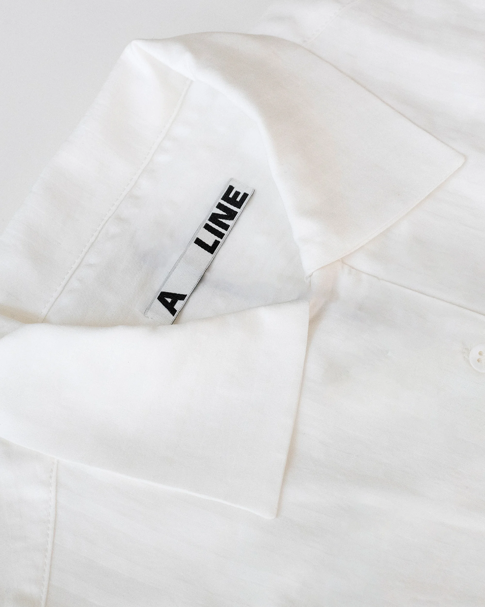 Close-up of a white button-down shirt with a collar and a label with black text, captured in a bright, neutral studio setting.