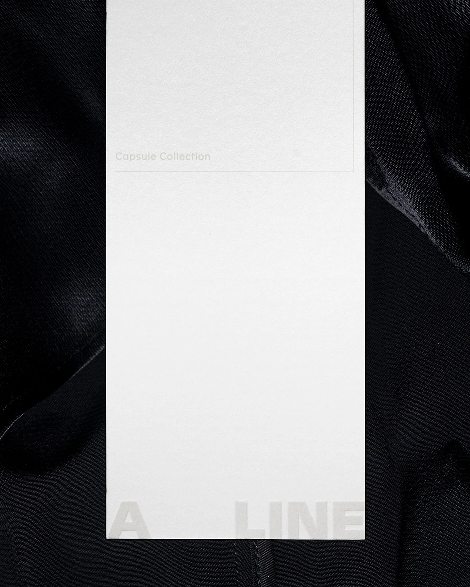 Close-up of a white tag with 'Capsule Collection' on dark fabric. Minimalist aesthetic with soft lighting.