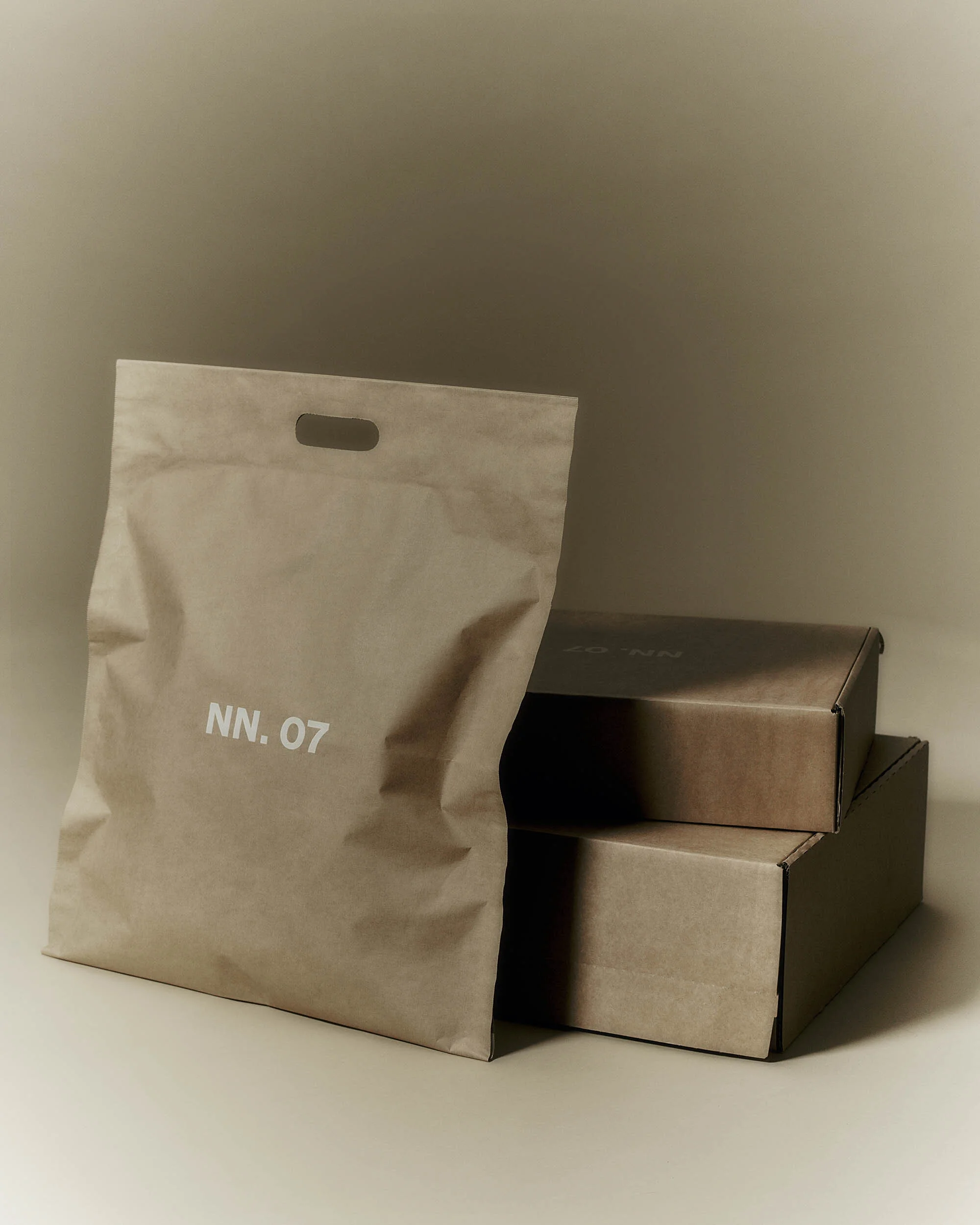 Minimalist product shot: kraft paper shopping bag with 'NN. 07' text next to stacked cardboard boxes, neutral background, soft lighting.