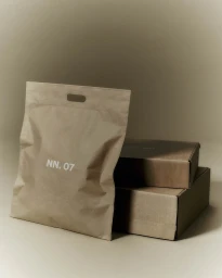 Brown paper shopping bag with white logo next to stacked cardboard boxes against a neutral studio backdrop.