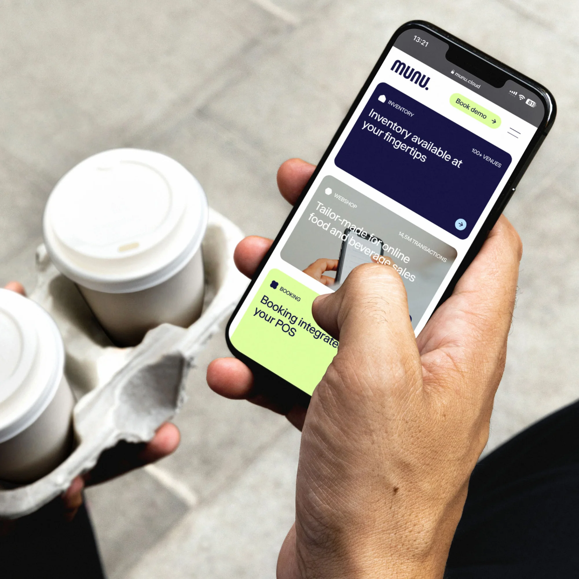 Hand holds a smartphone displaying a food and beverage sales app, with two white coffee cups in a cardboard carrier in the background.