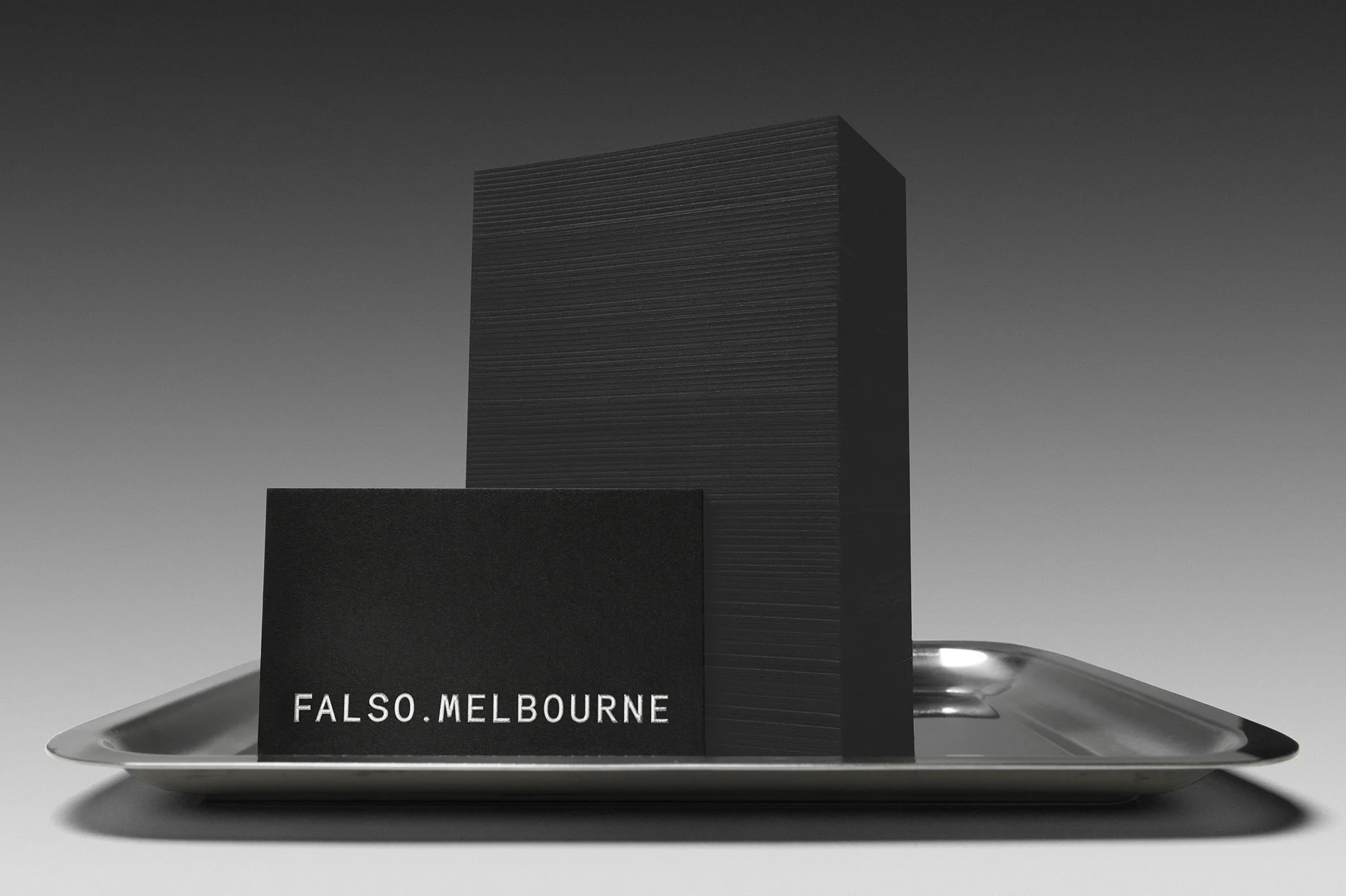 Stack of black business cards on a silver tray against a gray background. The front card has the text 'Falso.Melbourne'.