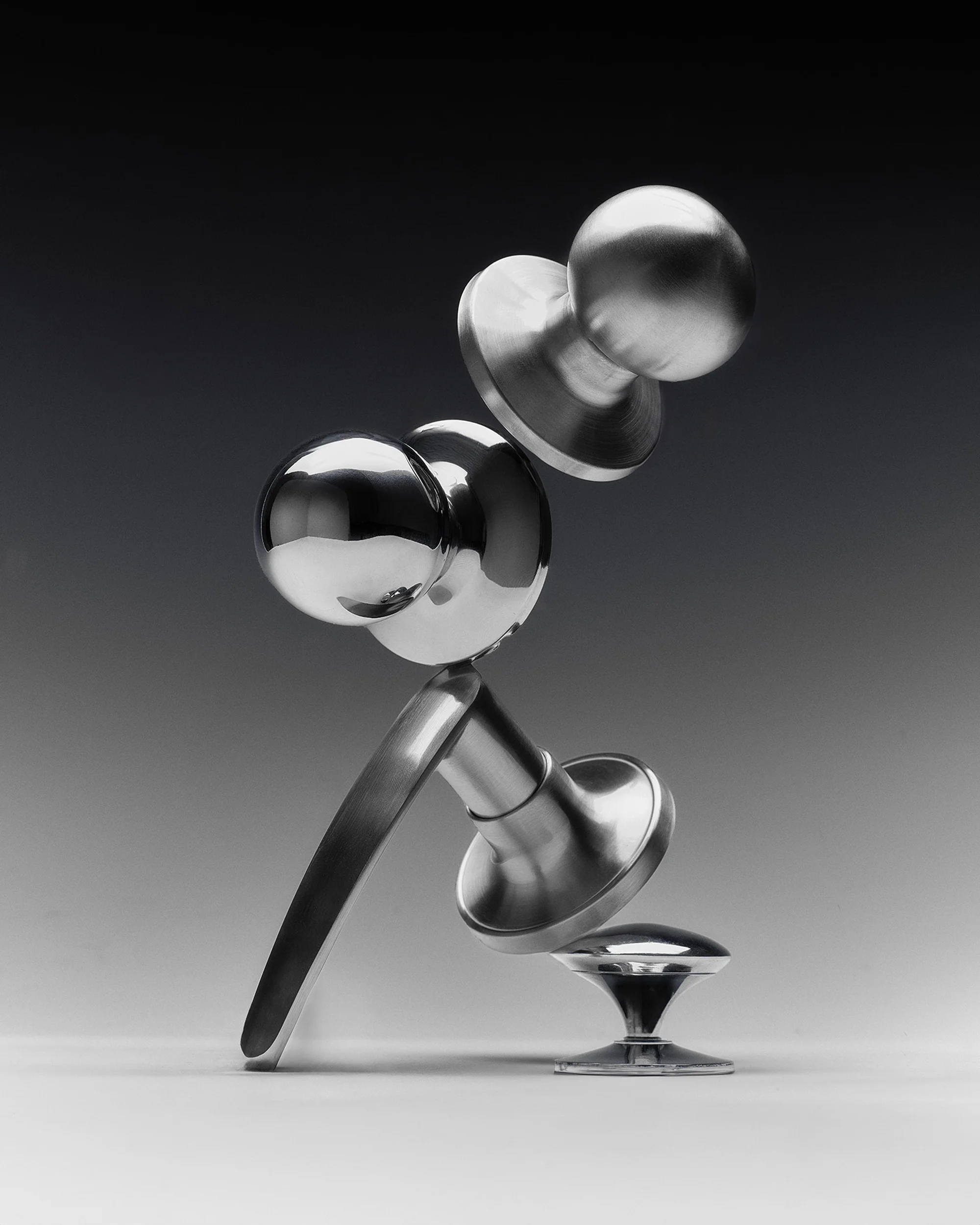Still life of chrome objects including spheres and pedestals arranged on a white surface against a dark grey gradient background.