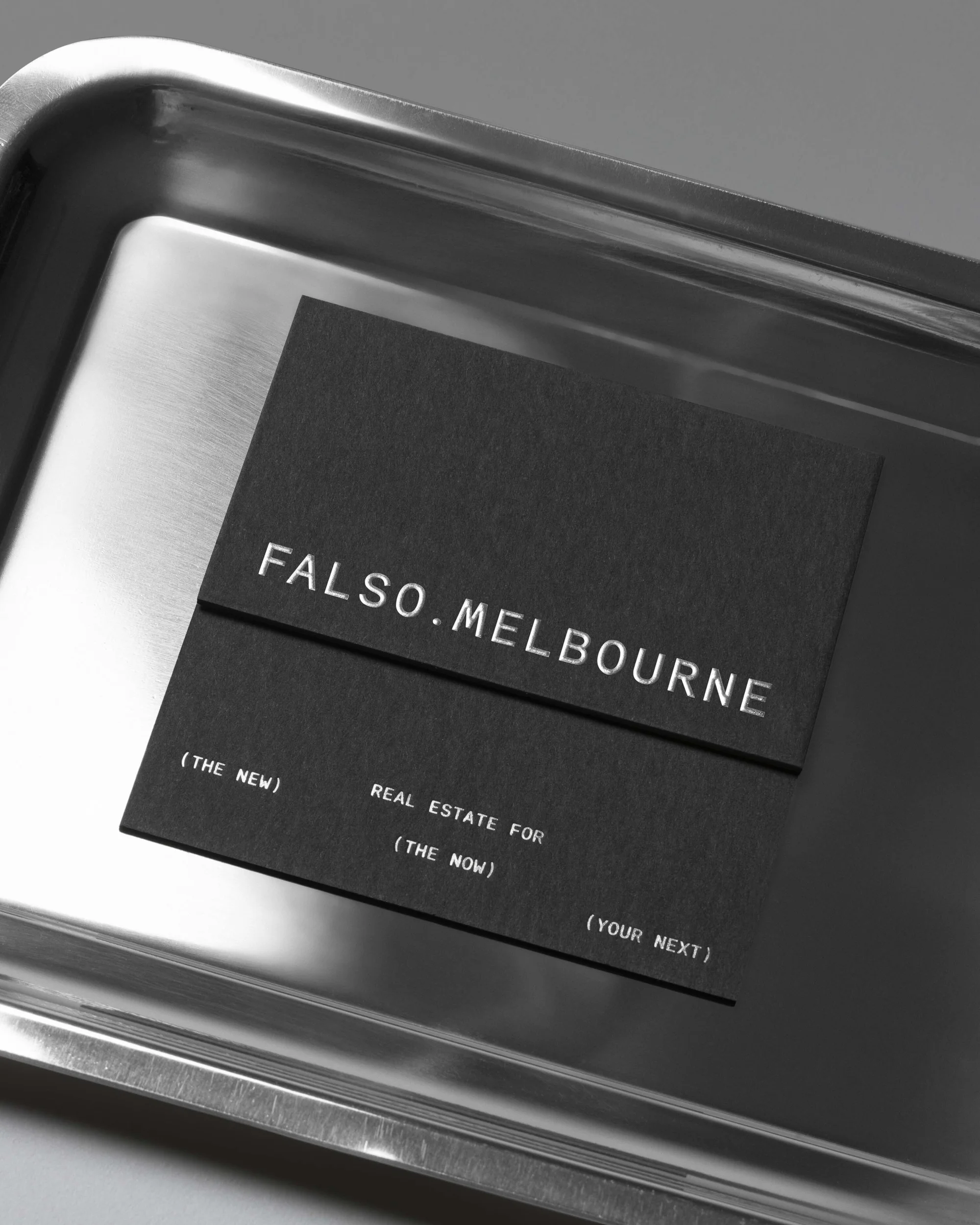 Close-up of a black business card on a metal tray with 'Falso.Melbourne' and real estate business details in silver text.