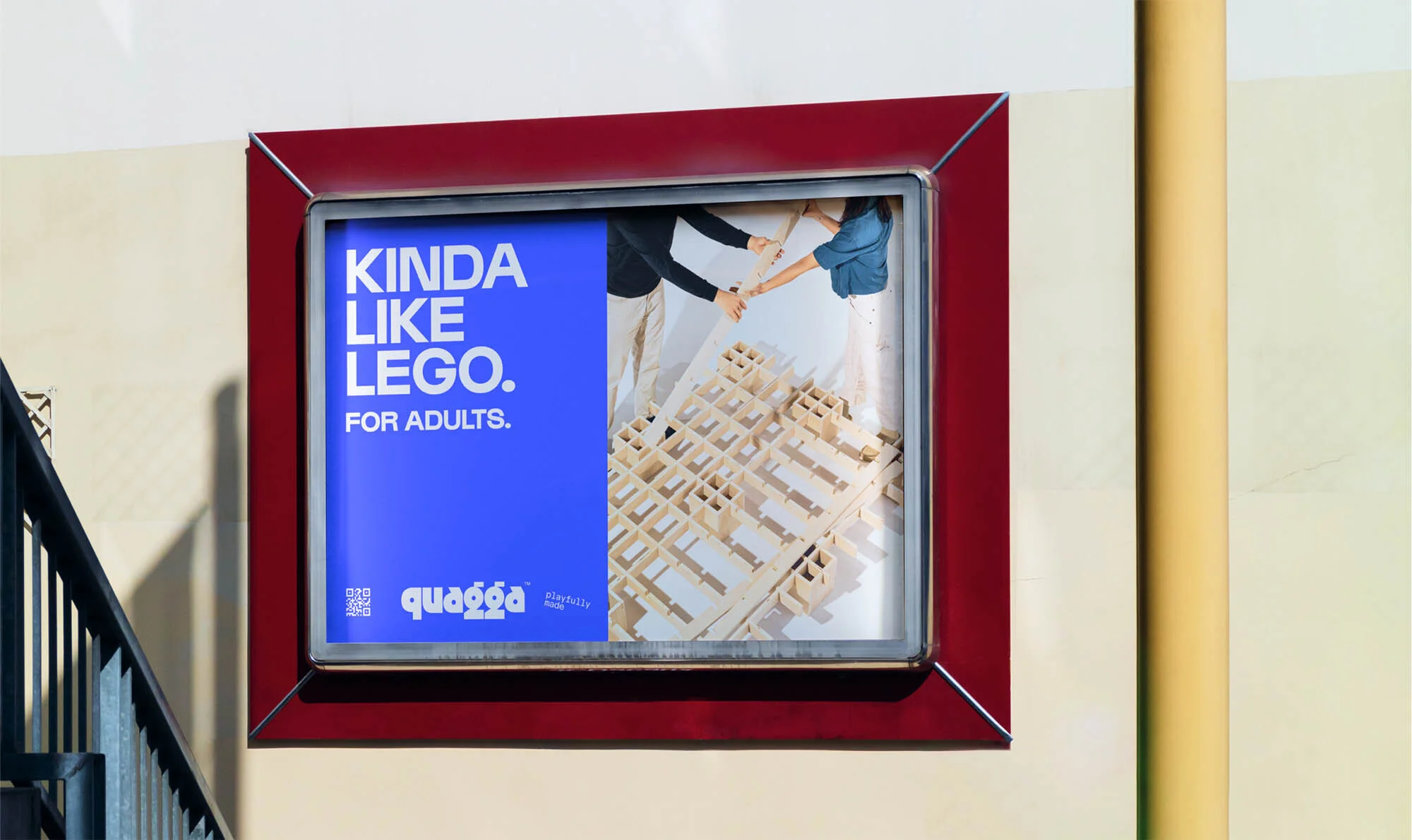 Framed advertisement for Quagga, a building toy. Two adults assemble the toy. Text reads: Kinda Like Lego. For Adults.