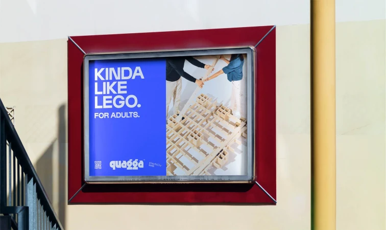 Framed advertisement for Quagga, a building toy. Two adults assemble the toy. Text reads: Kinda Like Lego. For Adults.