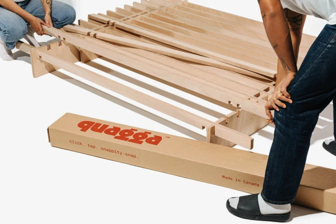 Two people assemble a light-wood bed frame from a cardboard box in a brightly lit photo studio with a clean, minimalist aesthetic.
