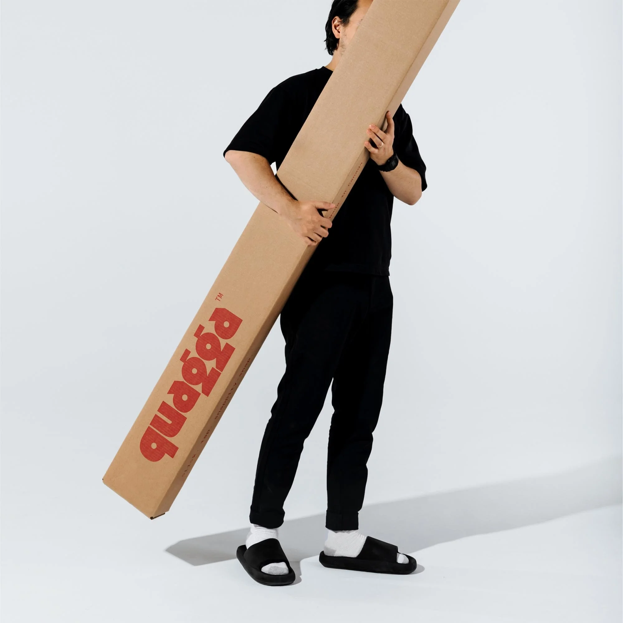 Adult male carries a large cardboard box labeled 'quagga' in red. He is dressed in casual black clothing, including slides, against a plain white background.