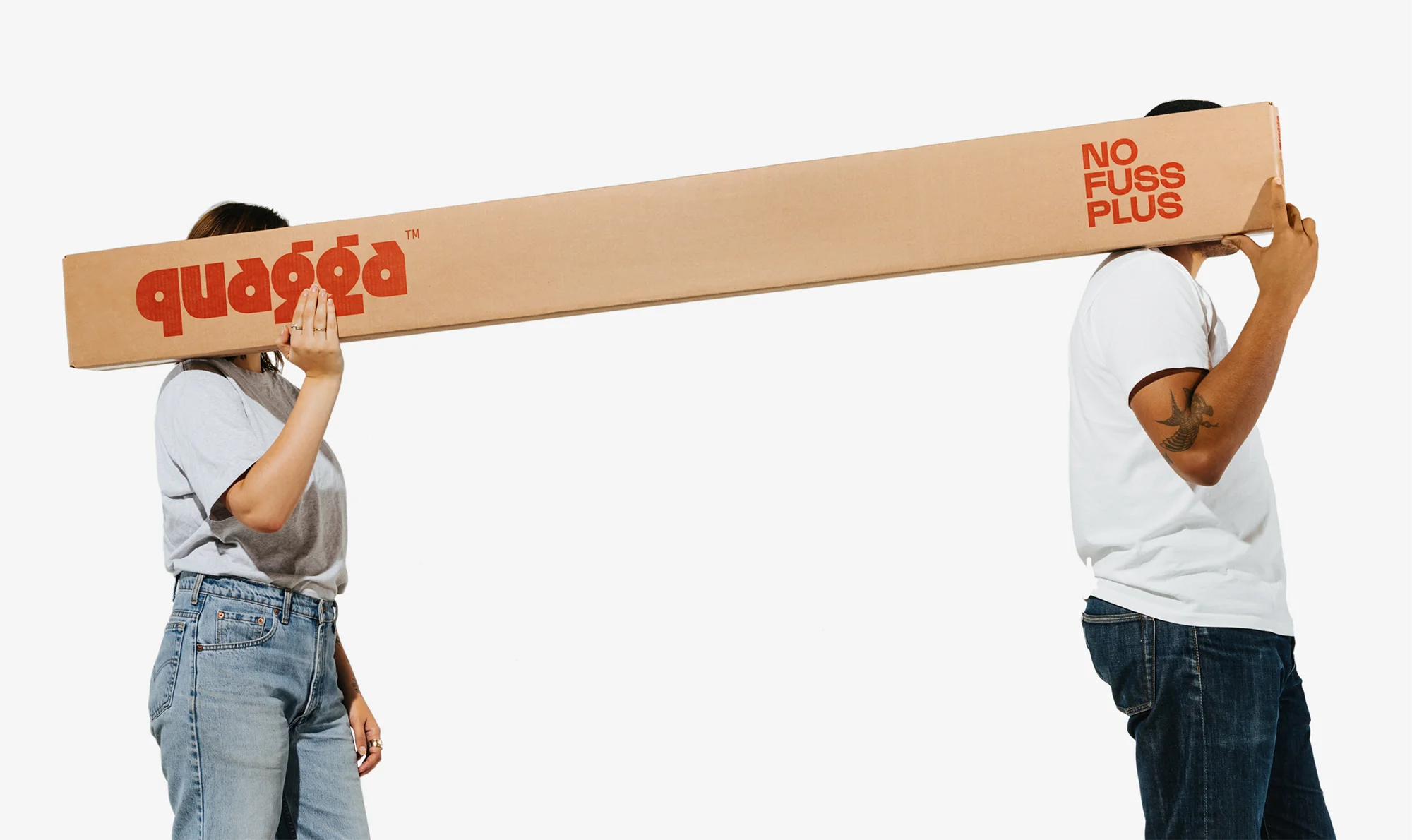 Woman and man carrying a large cardboard box with red text on a white background in a studio setting, facing away from each other.