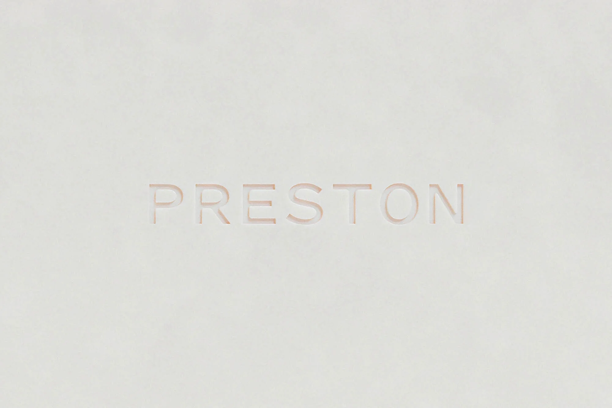 The word PRESTON is carved into a light beige textured surface, set against a creamy, monochrome background. The font is sans-serif and modern.