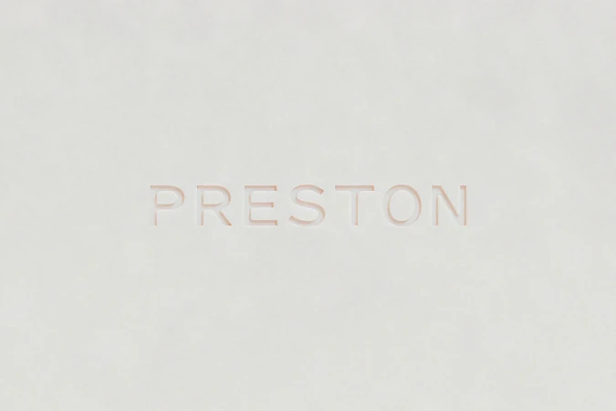 The word PRESTON is carved into a light beige textured surface, set against a creamy, monochrome background. The font is sans-serif and modern.