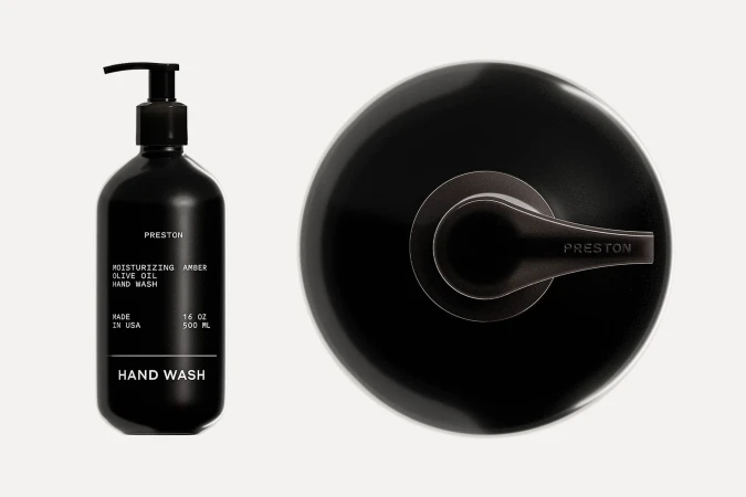 A black Preston hand wash bottle with a pump dispenser sits next to a modern, minimalist black metal shower faucet on an off-white surface.