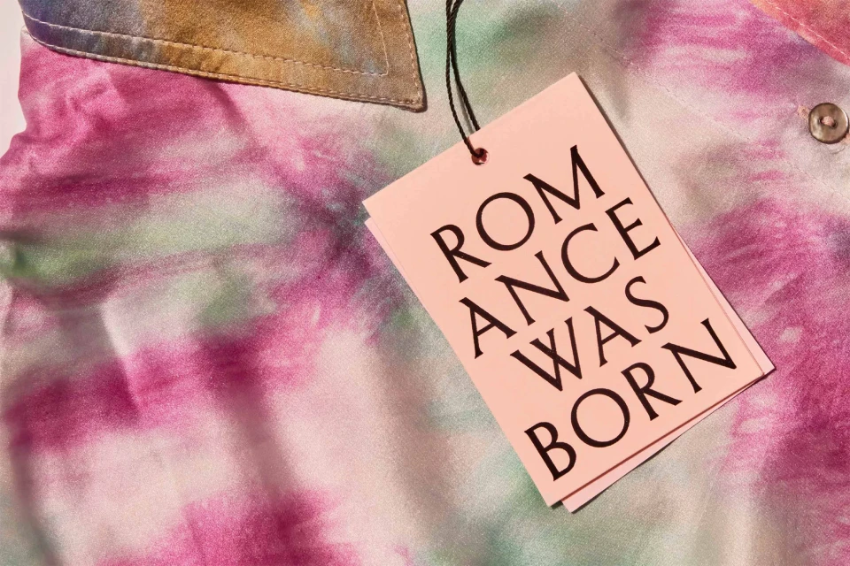 Close-up of a pink and green tie-dye shirt with a paper tag that reads, 'Romance Was Born' attached with a black string.