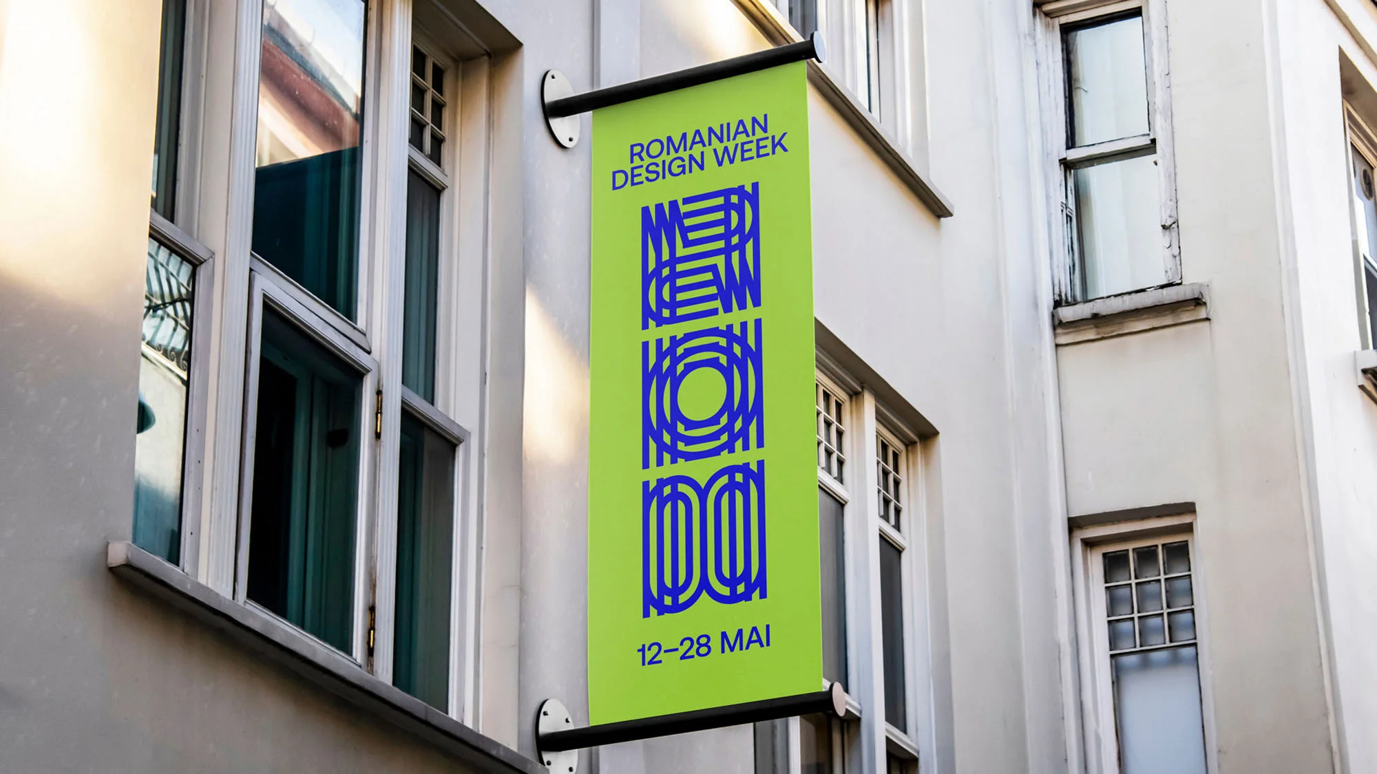 A green vertical banner for Romanian Design Week hangs on a building with white-framed windows. The banner features blue text and graphics.
