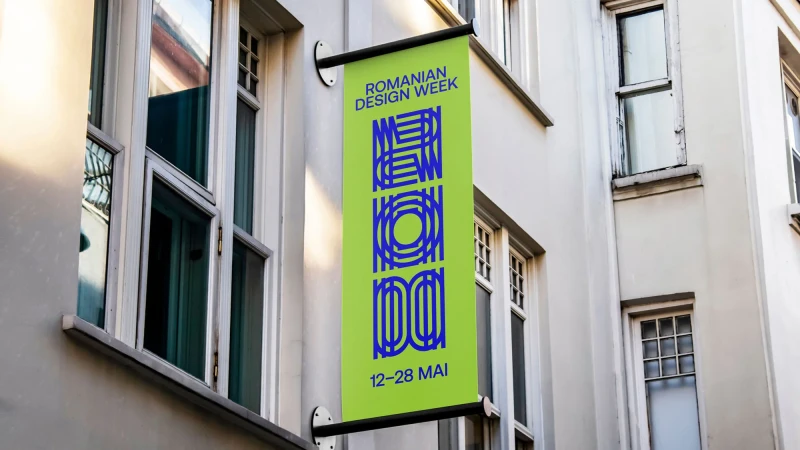 A green vertical banner for Romanian Design Week hangs on a building with white-framed windows. The banner features blue text and graphics.