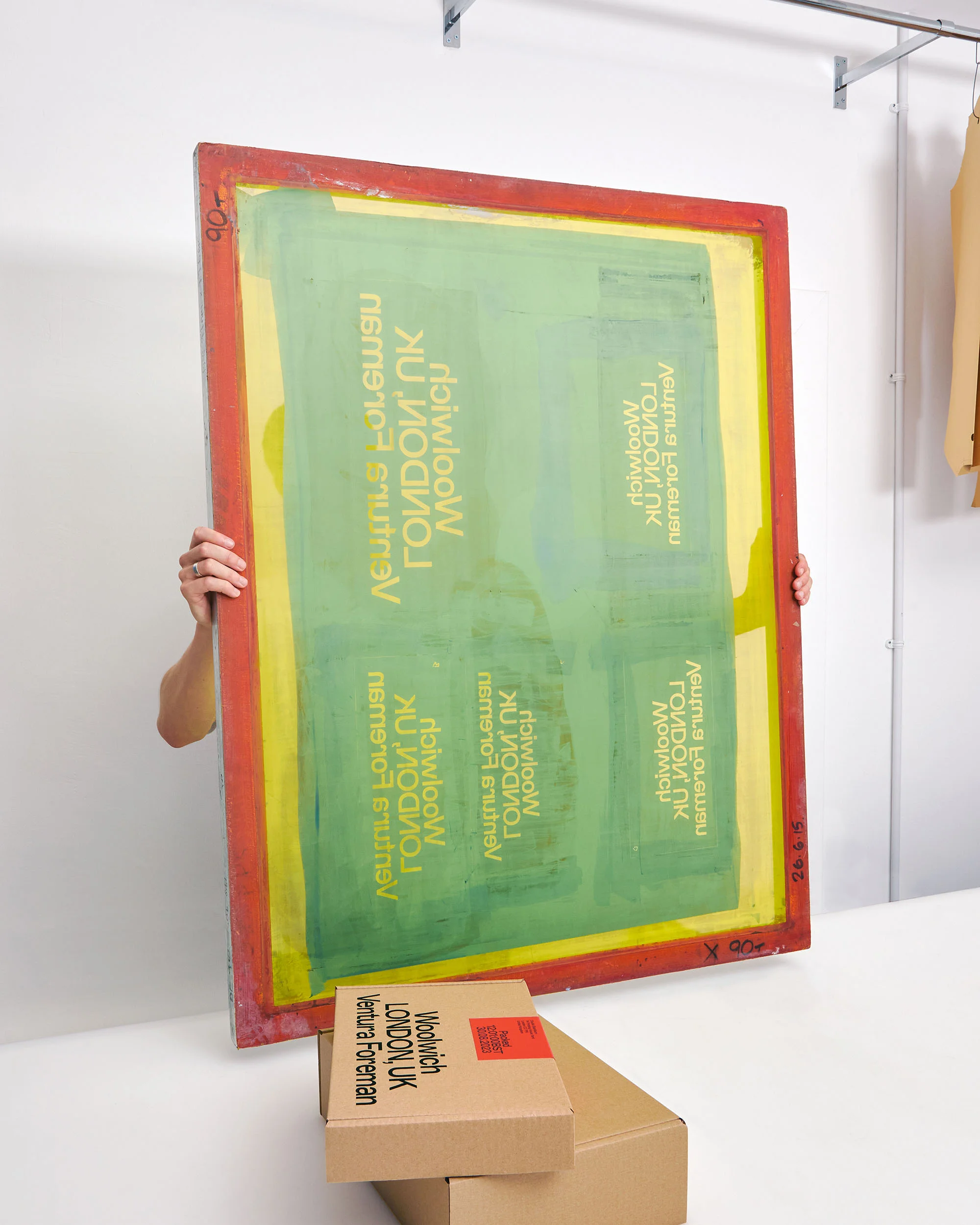Person holding a large silkscreen with red frame and green and yellow design, with cardboard boxes stacked below in a studio.