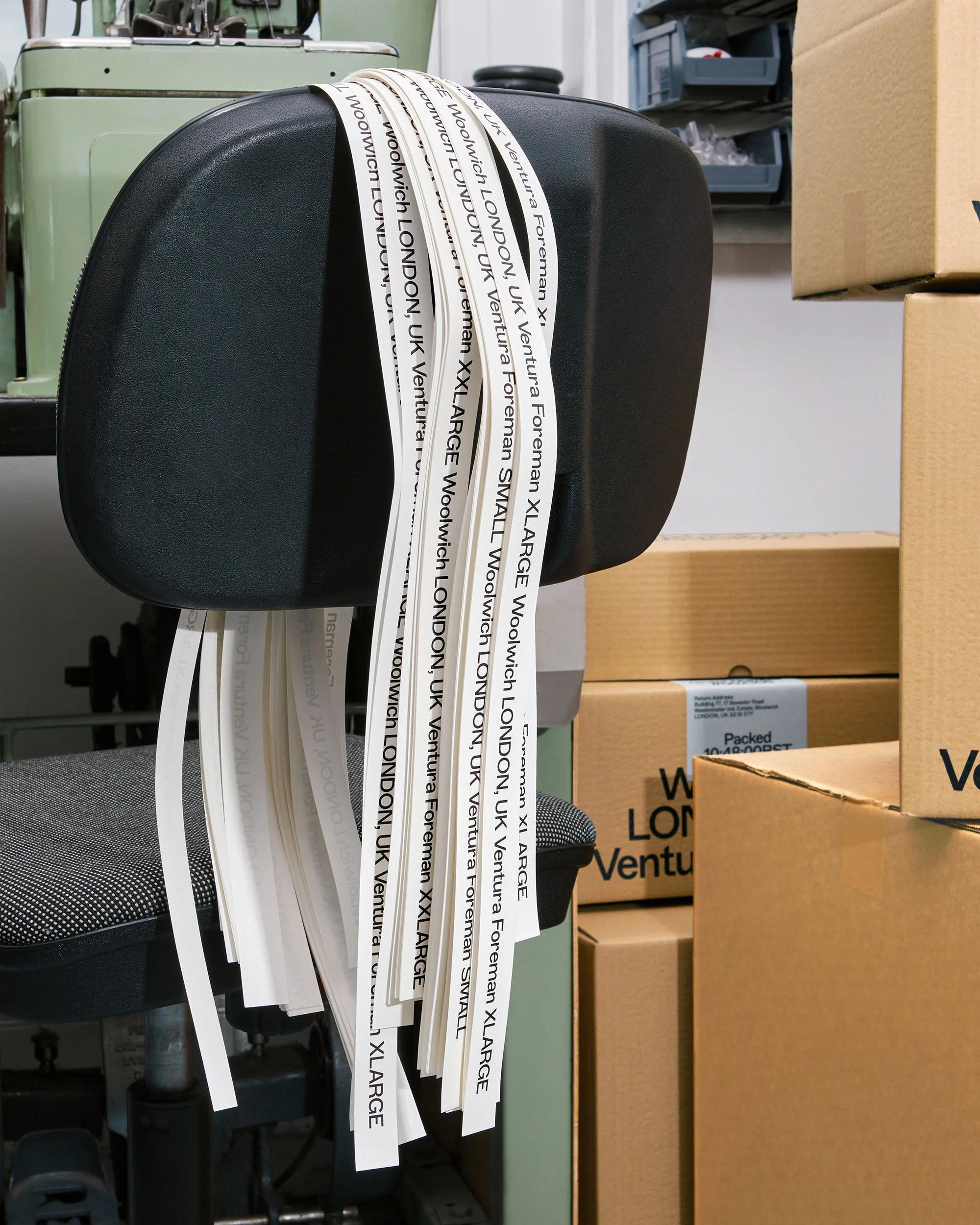 Black office chair with labels listing London locations and size variations. Boxes in background, sewing machine to the left.