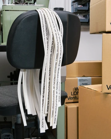 Black office chair with labels listing London locations and size variations. Boxes in background, sewing machine to the left.