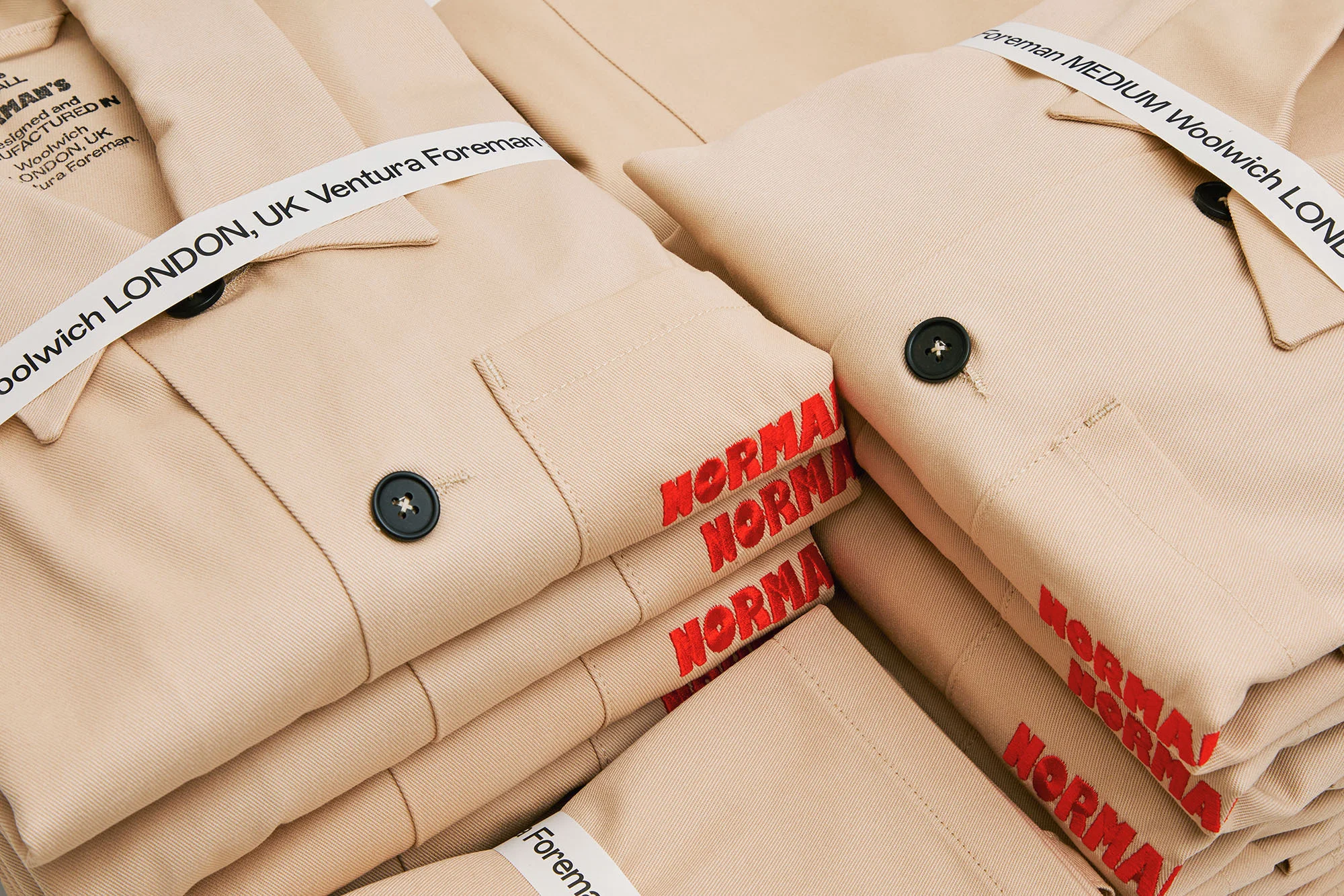 Pile of beige Norman Woolwich London UK Ventura Foreman shirts with black buttons and red text detail, offering a minimalist, clean aesthetic.