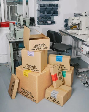 Stacked cardboard shipping boxes branded with text in a workshop setting with sewing equipment and a chair.