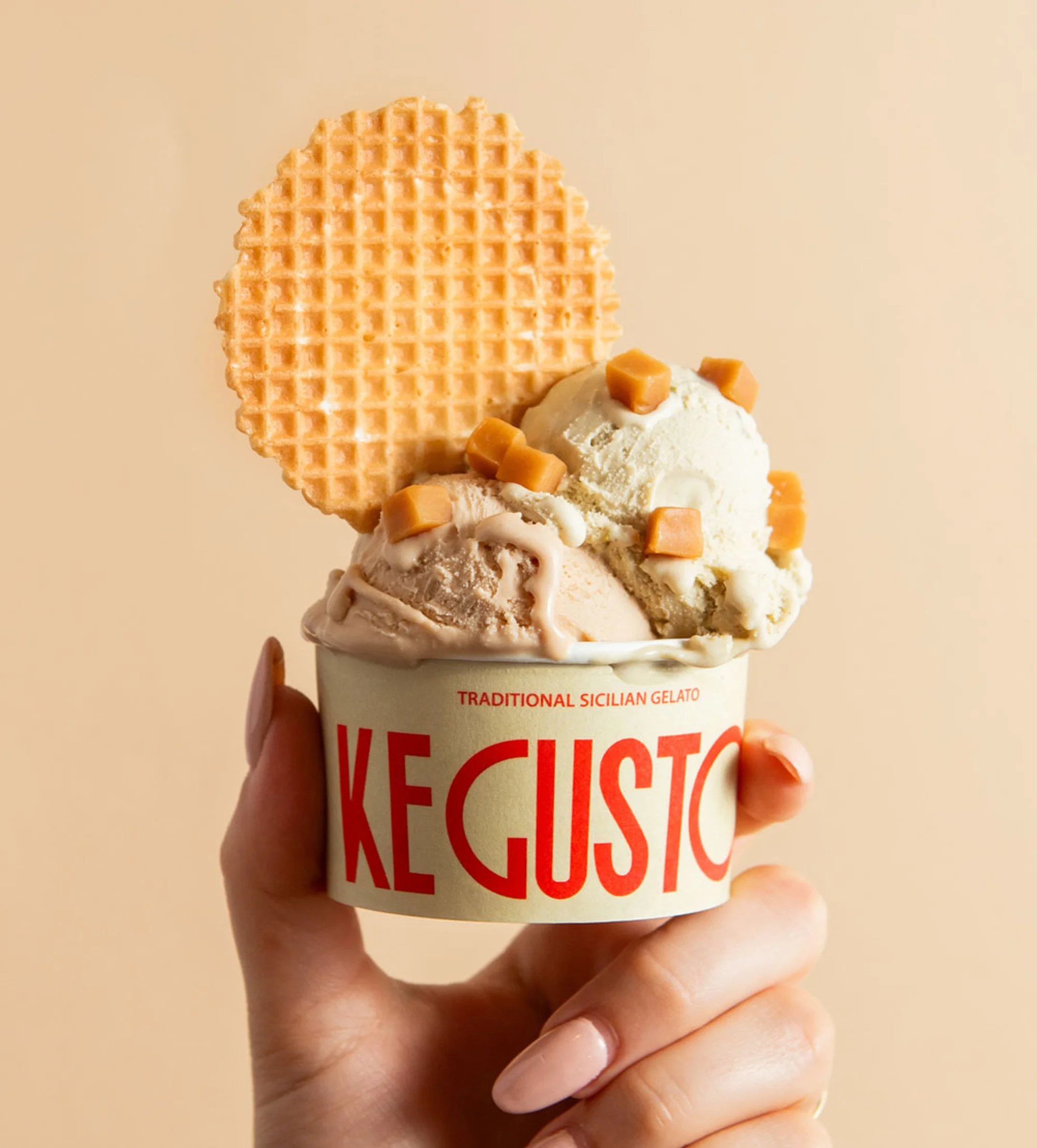 A hand holds a paper cup filled with gelato, topped with caramel pieces and a waffle cookie, set against a neutral backdrop.