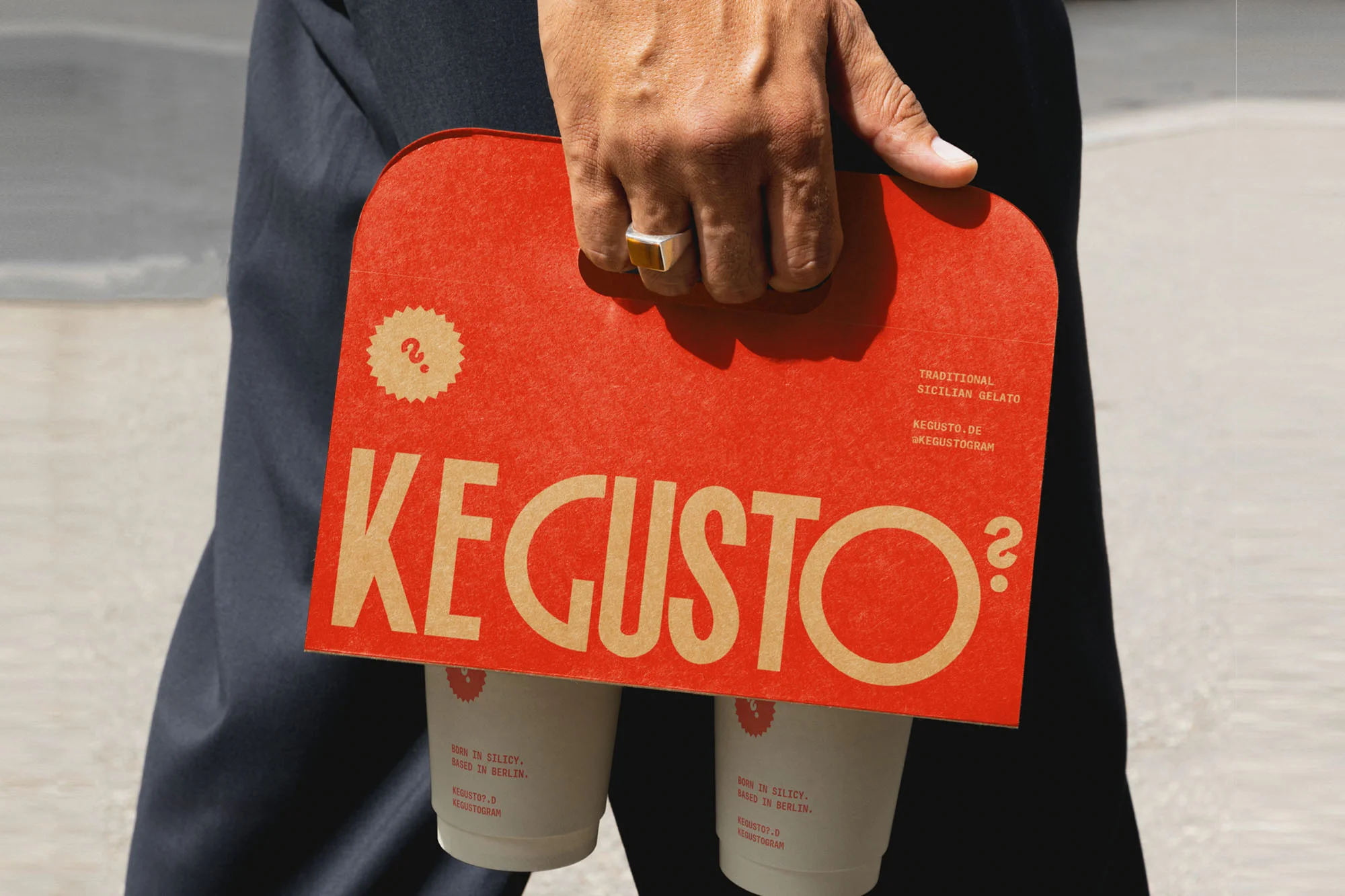 A person holds a red cardboard carrier with two cups of KE GUSTO gelato on a bright day, showcasing the brand's logo and product.