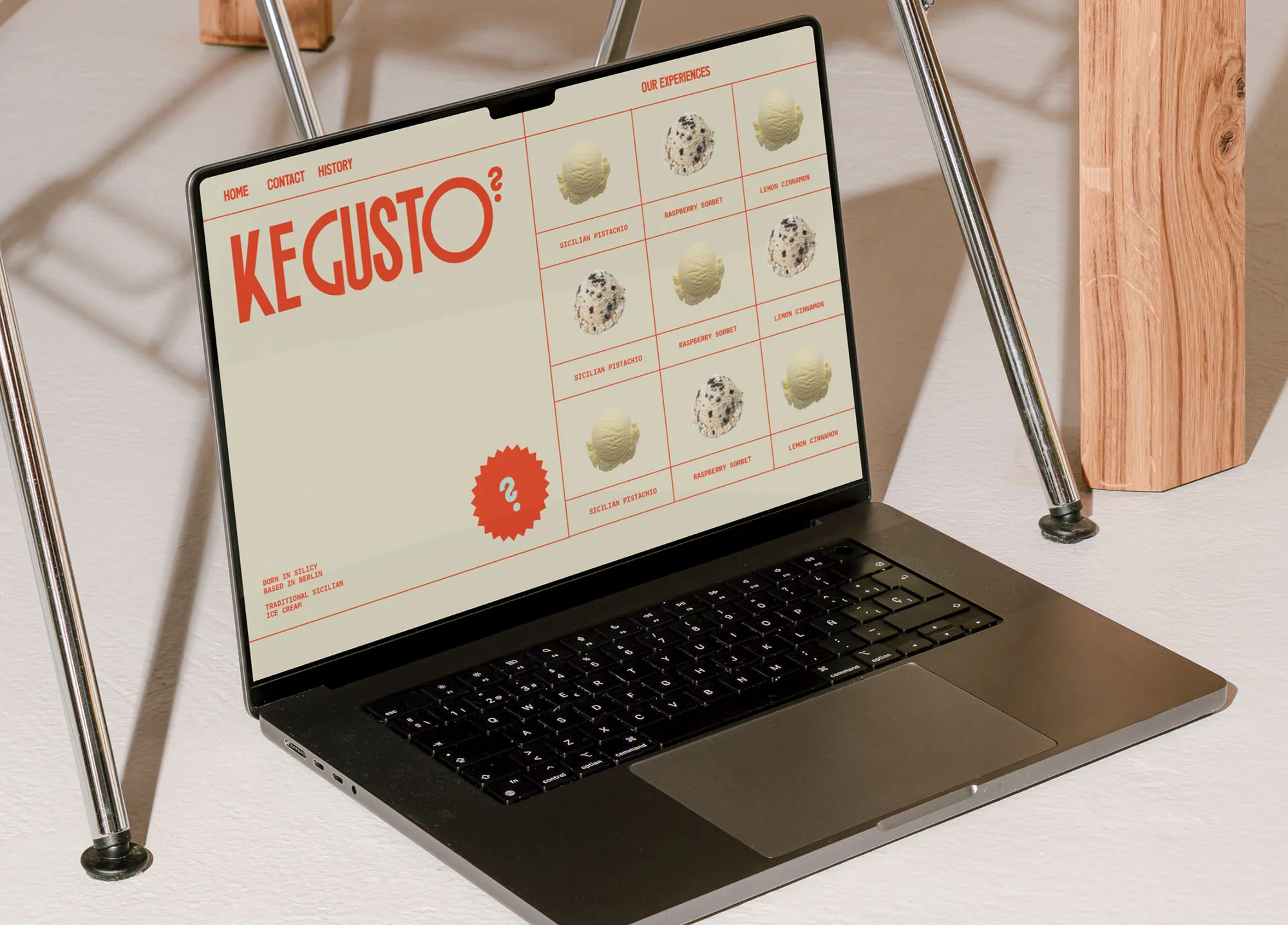 Open laptop displays KE GUSTO webpage with ice cream flavors; placed on white table with chair legs and wood accent.