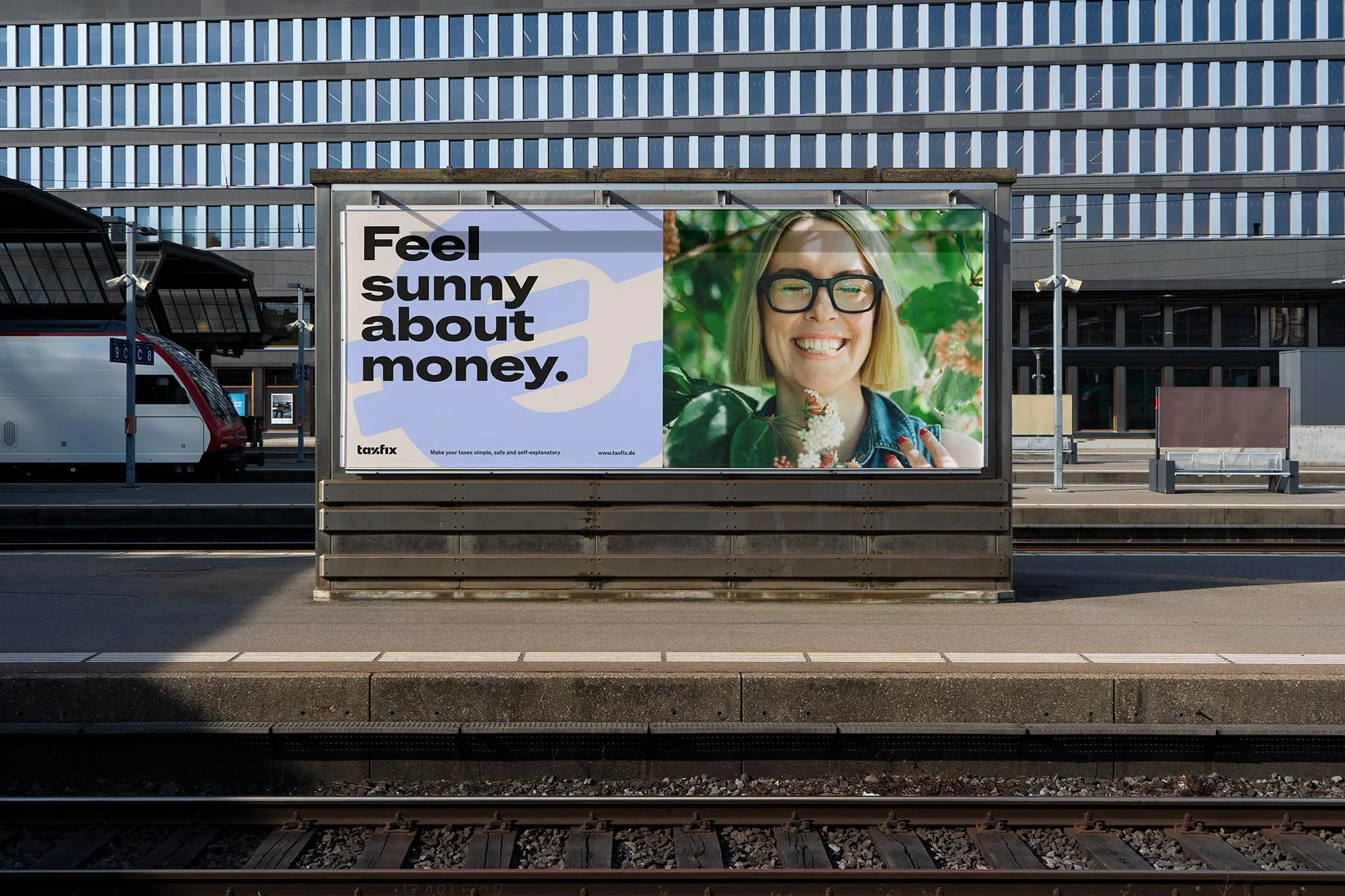 Outdoor advertising billboard featuring a smiling woman in glasses, placed at a train station with a passenger train visible behind.