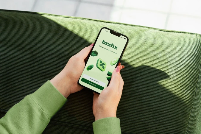 Woman holds a smartphone displaying a Taxfix app interface with floating graphic elements, against a green couch background.