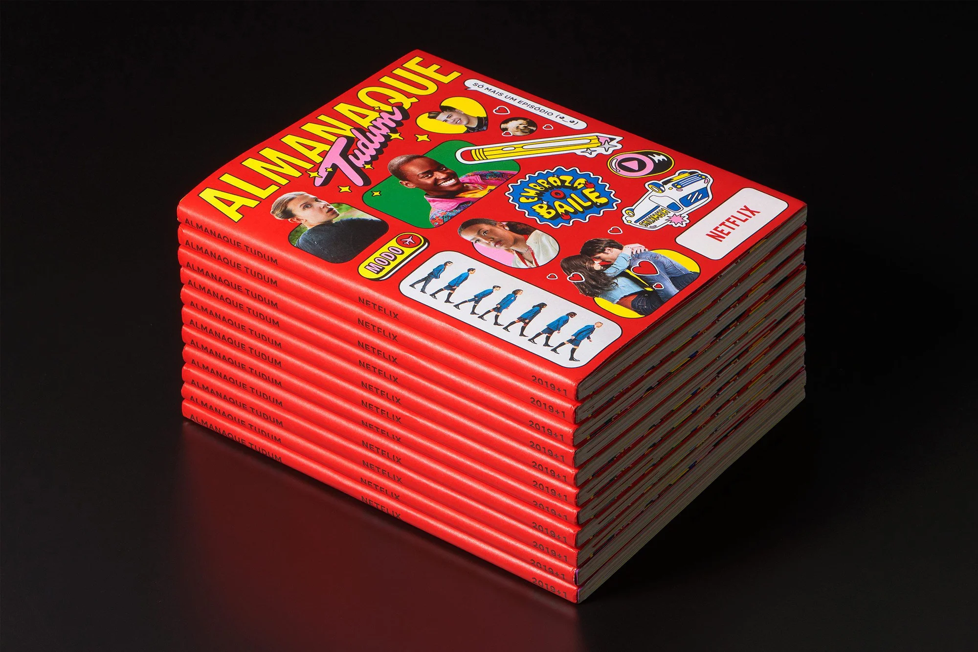 Stack of Almanaque Tudum magazines on a black surface, featuring diverse faces and sticker-like graphics on the red covers.