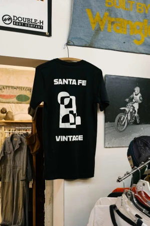 Black graphic tee hanging indoors on a white wall. Tee reads 'Santa Fe Vintage.' Vintage graphic apparel in a retail environment.