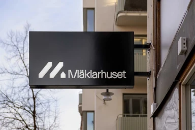 Black rectangular sign with white Maklarhuset logo mounted on a beige building exterior with bare trees in background