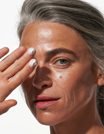 Close-up of a mature woman applying cream to her face, showcasing skincare and beauty with natural gray hair and clear skin.