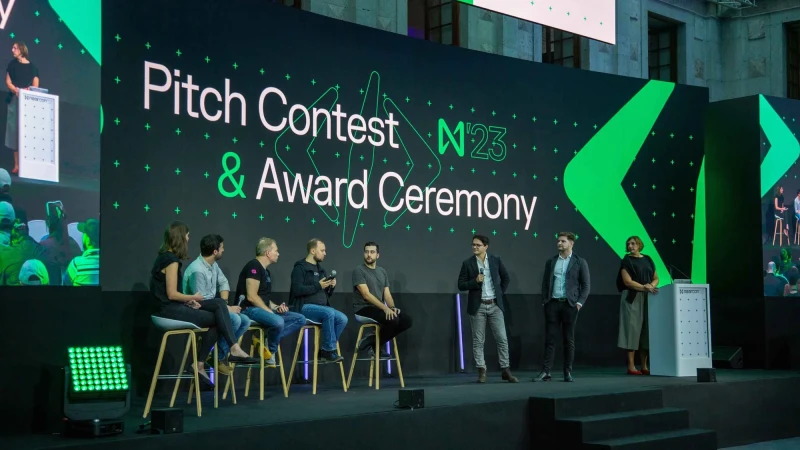 Panel discussion at a conference with a group of people on stage sitting on stools in front of a large screen displaying text and graphics.