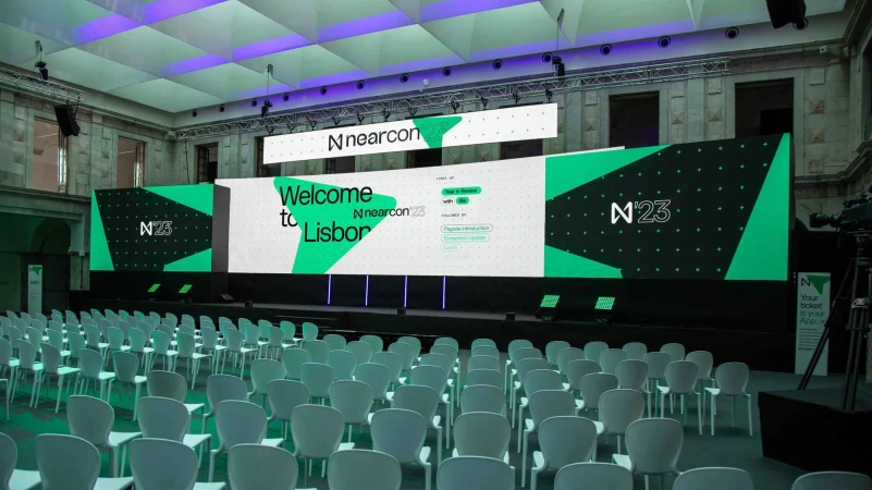 Auditorium stage with large LED screens displaying event branding, rows of empty white chairs, and conference setup.
