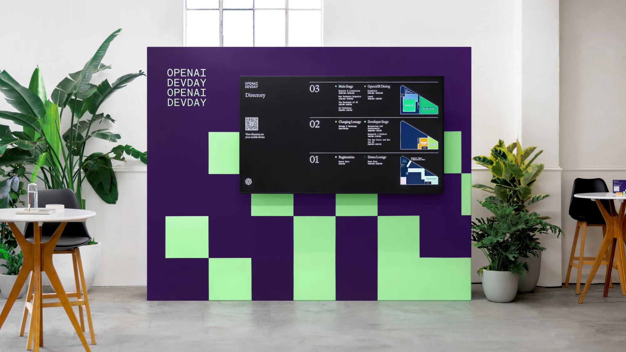OpenAI DevDay event directory on a screen with schedule information, located in an indoor event space with plants and modern furniture.