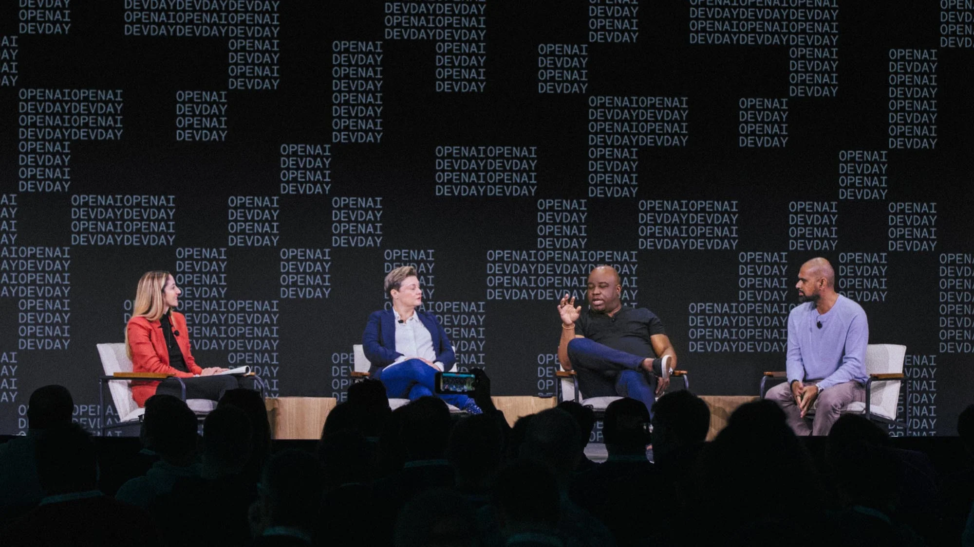 Four panelists sitting on stage having a discussion in front of an audience, with a backdrop featuring repeating text.