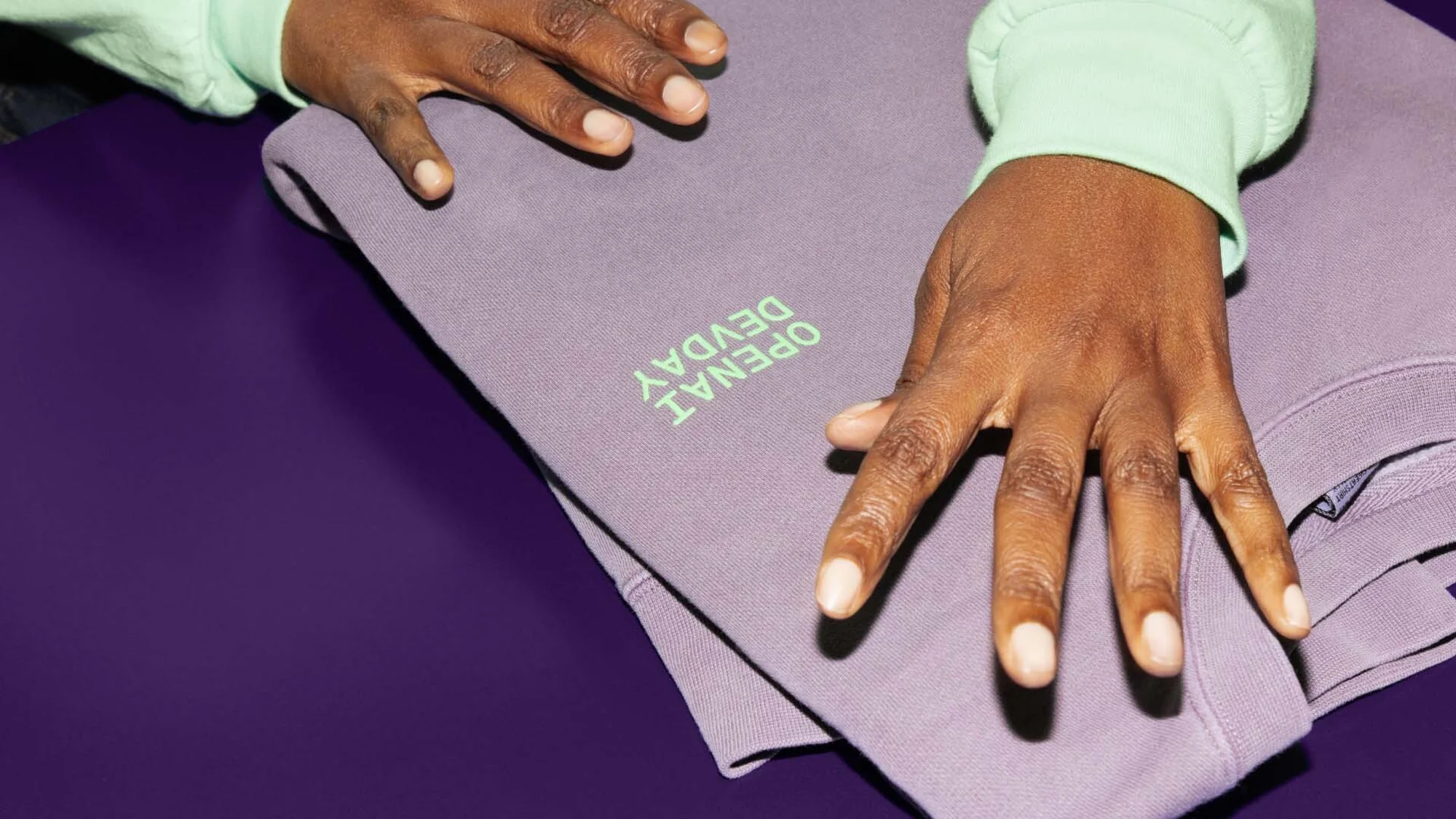 Folded purple sweatshirt with light green embroidered text on dark purple background, styled with brown skinned hands and light green sleeves.